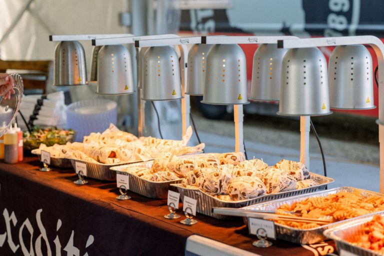 Self-serve catering buffet spread with brats and sides under daylight.