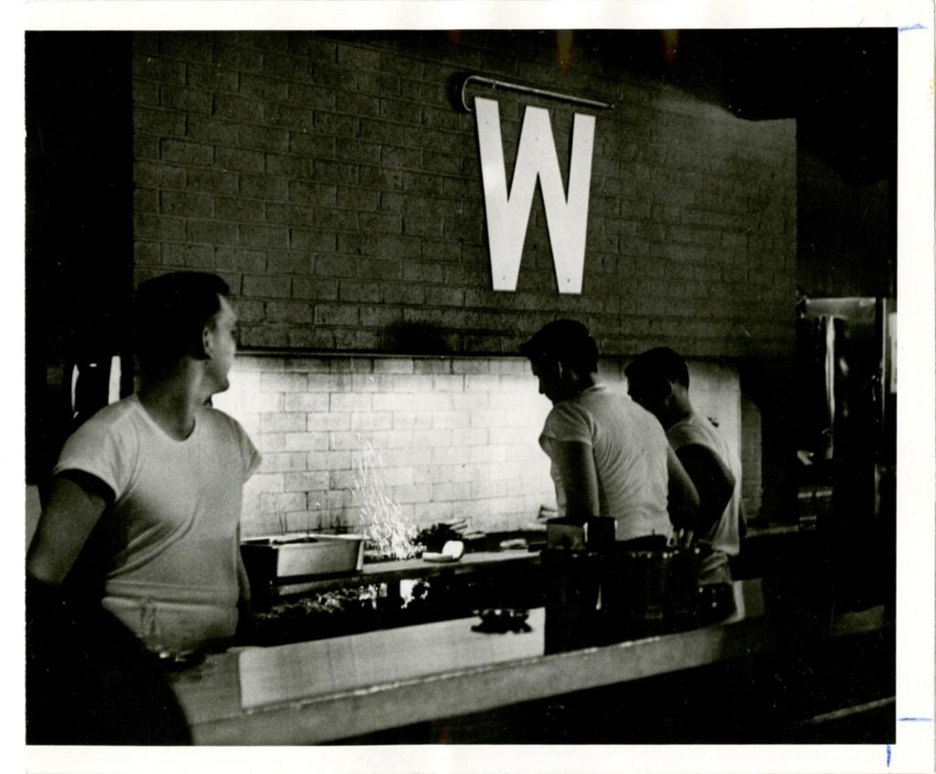Young State Street Brats grill cooks in white tees working the counter under a large white W banner, in black and white.